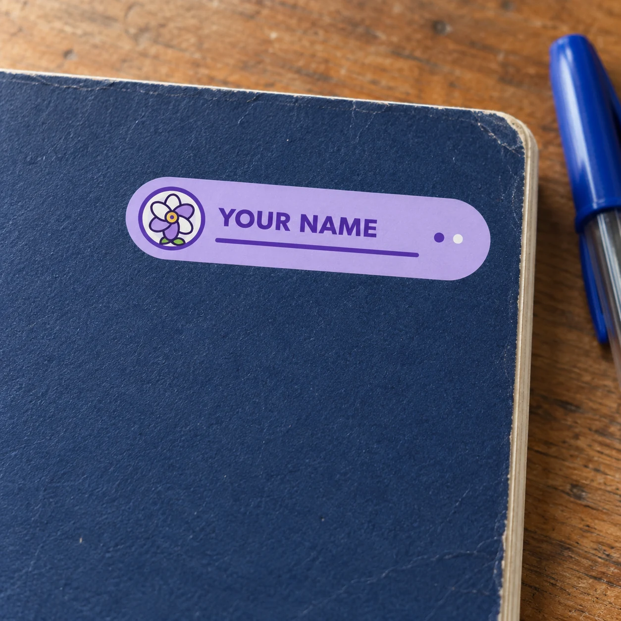 Lavender flower name tag sticker reading YOUR NAME on a blue exercise book cover