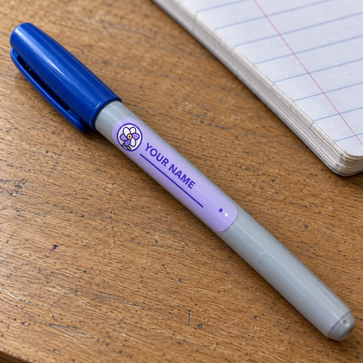 Lavender flower name tag sticker reading YOUR NAME applied lengthwise to a blue-capped pen